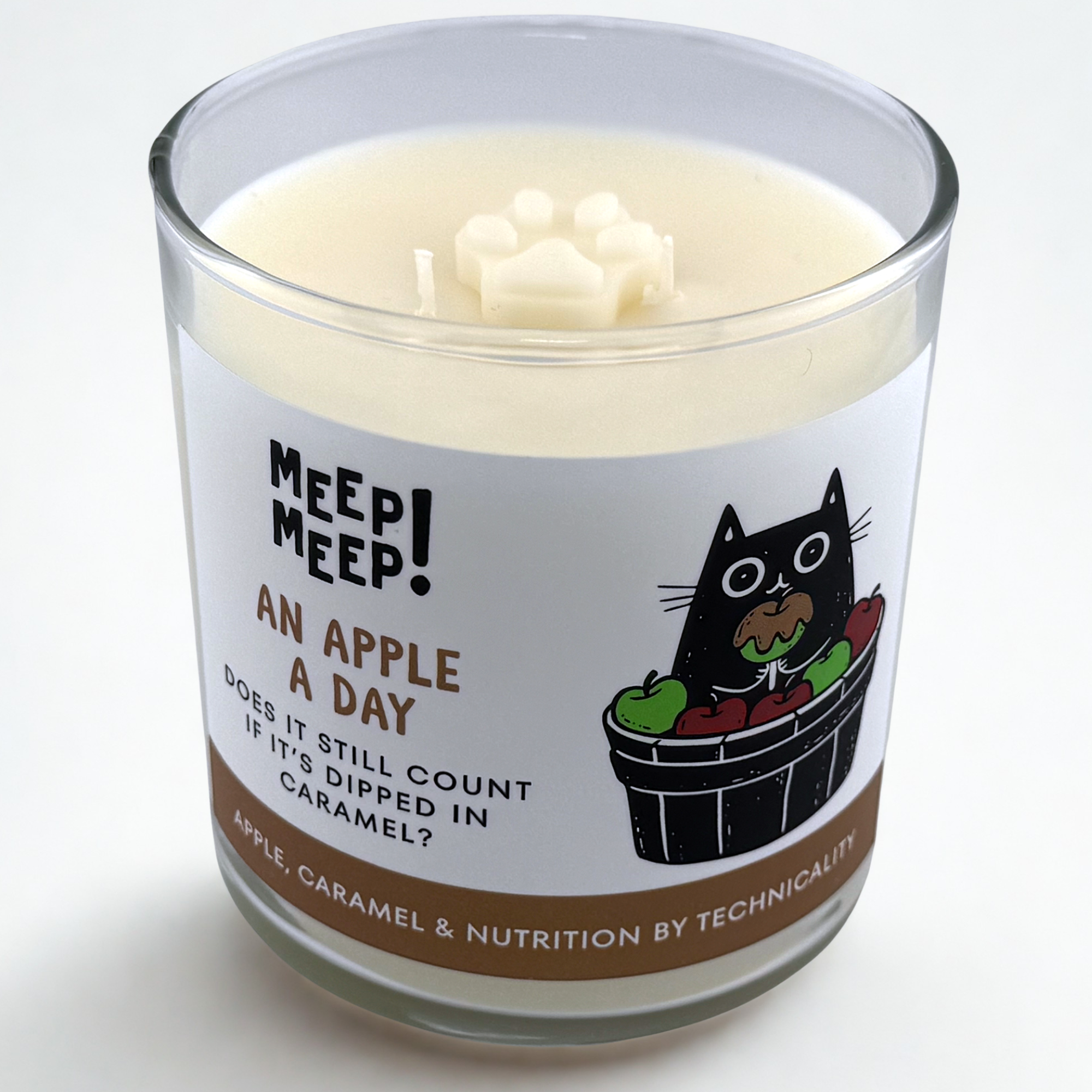 Photograph of Meep Meep candle with wax pawprint embed on top. Label has an Illustration of a cat holding a caramel apple and sitting in a bucket full of red & green apples. Text on white background says "An Apple A Day. Dos It Still Count If It's Dipped In Caramel?"
Bottom of candle label reads Apple, Caramel & Nutrition by Technicality