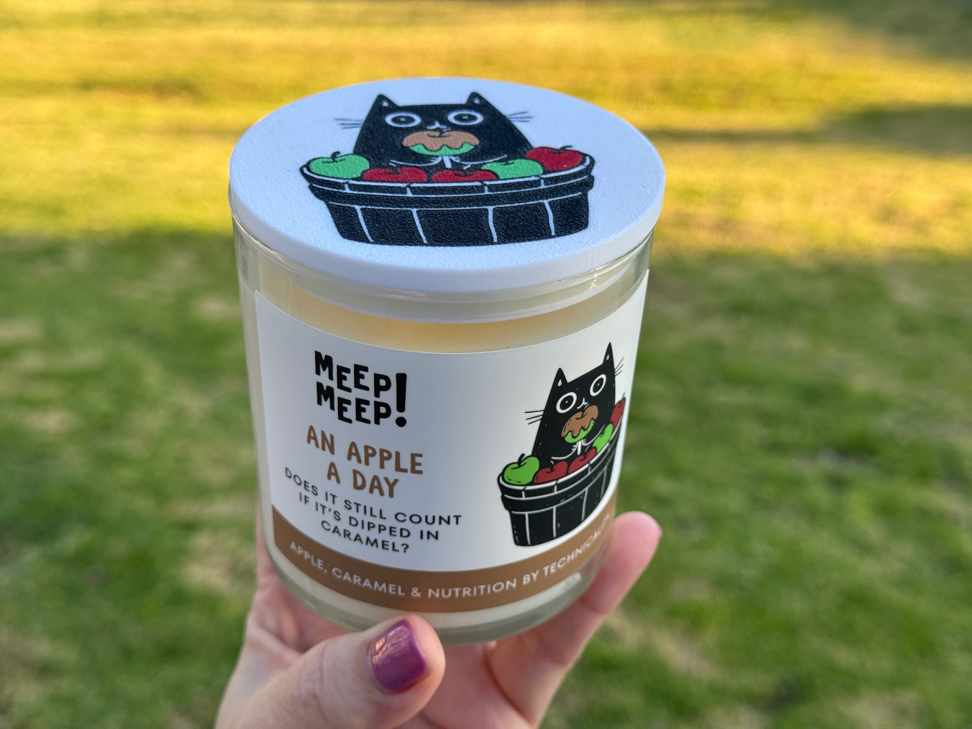 An Apple A Day candle being held in a hand. The candle has an illustration of a Black Cat sitting in a barrel of apples and holding a caramel apple. Text on candle reads: An Apple A ay. Does it still count if it's dipped in caramel? Apple, Caramel & Nutrition by Technicality
