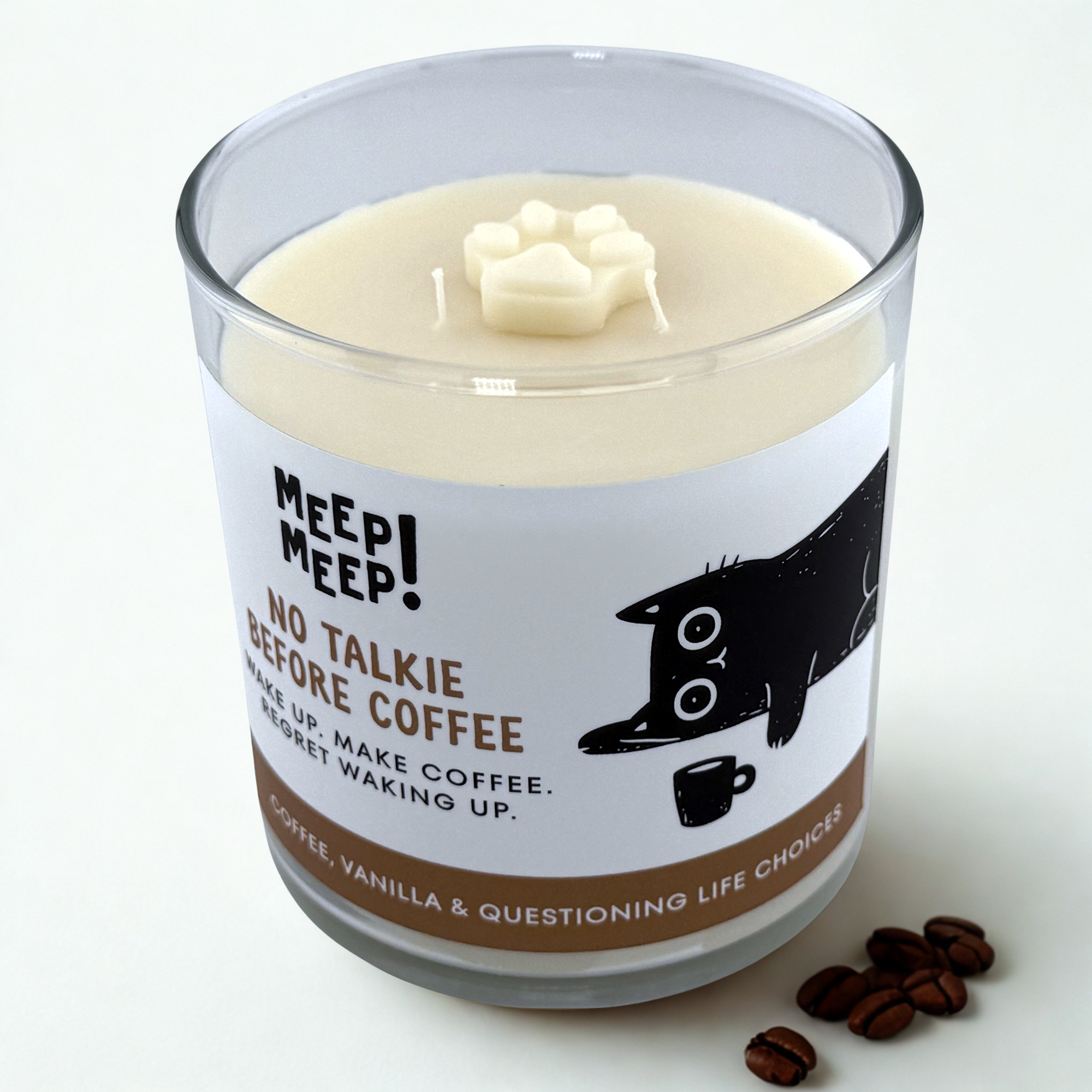 Photograph of Meep Meep candle in glass jar with wax paw print embed on top. Label has an Illustration of a black cat lying on the floor reaching for a coffee cup. Text on white background says "No TalkieBefore Coffee. wake up. make coffee.regret waking up."
Bottom of candle label reads coffee, vanilla & questioning life choices