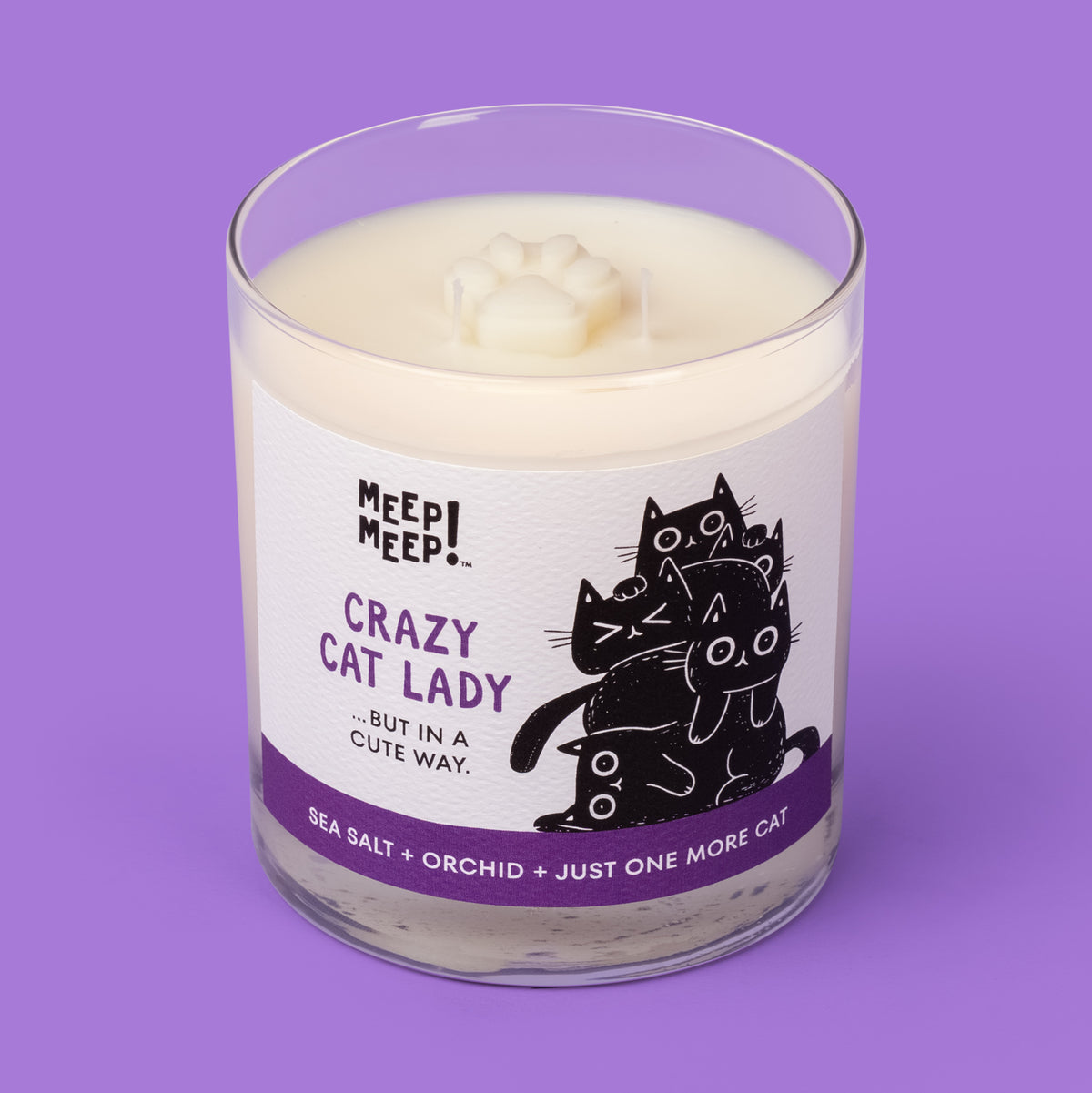 Meep Meep Crazy Cat Lady candle in a clear glass jar with a white and purple label showing a pile of black cats. Jar is angled so we can see the paw embed on top of candle.