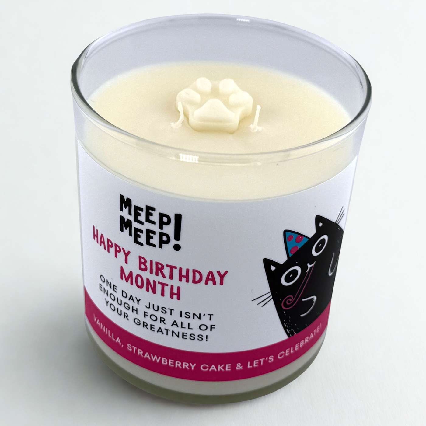 A photo of a glass Candle with label that has a title written in pink text, Happy Birthday Month. And more text that says, One Day just isn't enough for all of your greatness. Vanilla, Strawberry Cake & Let's celebrate. The cute black Meep Meep cat is on the right wearing a party hat and a party horn in it's mouth. 