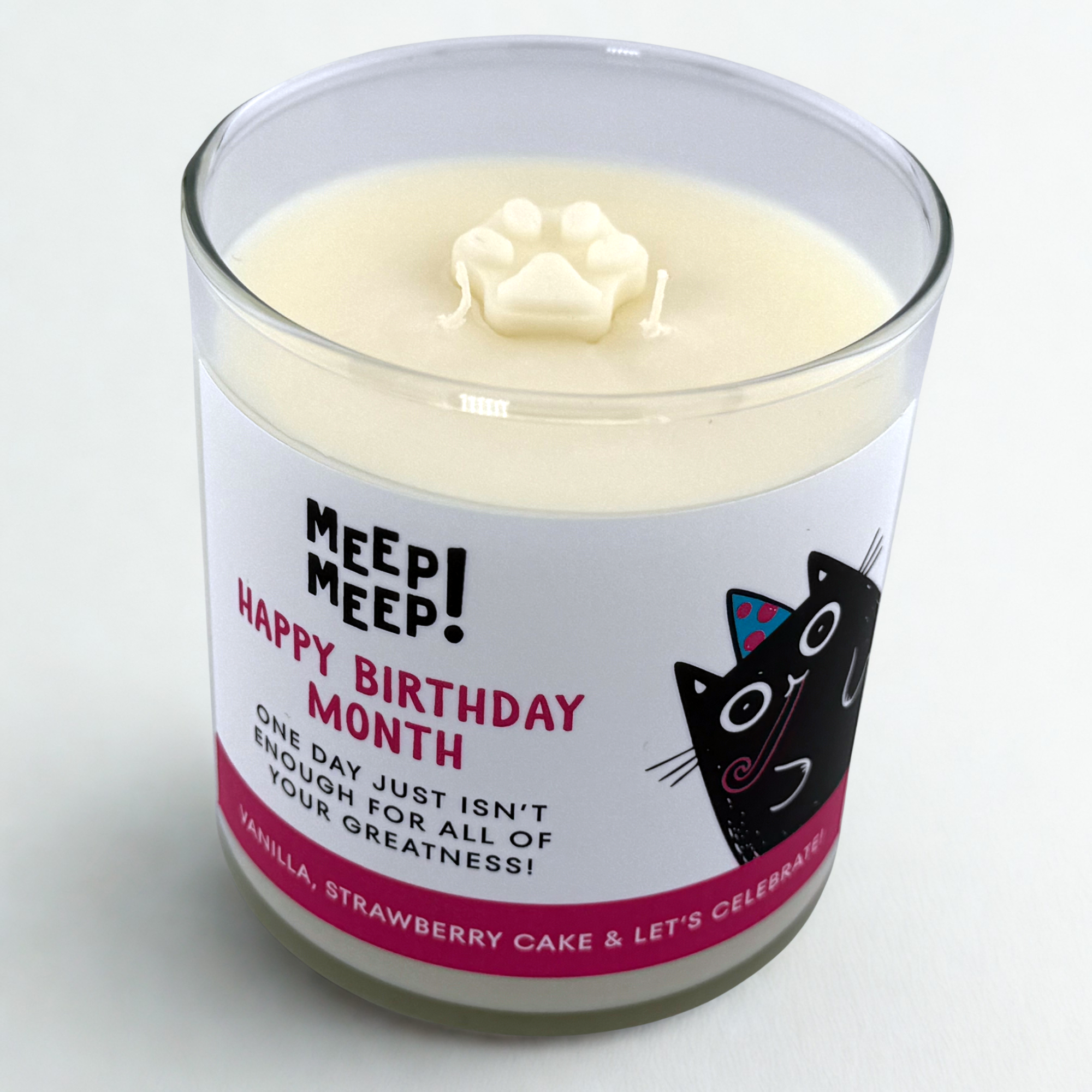 A photo of a glass Candle with label that has a title written in pink text, Happy Birthday Month. And more text that says, One Day just isn't enough for all of your greatness. Vanilla, Strawberry Cake & Let's celebrate. The cute black Meep Meep cat is on the right wearing a party hat and a party horn in it's mouth. 