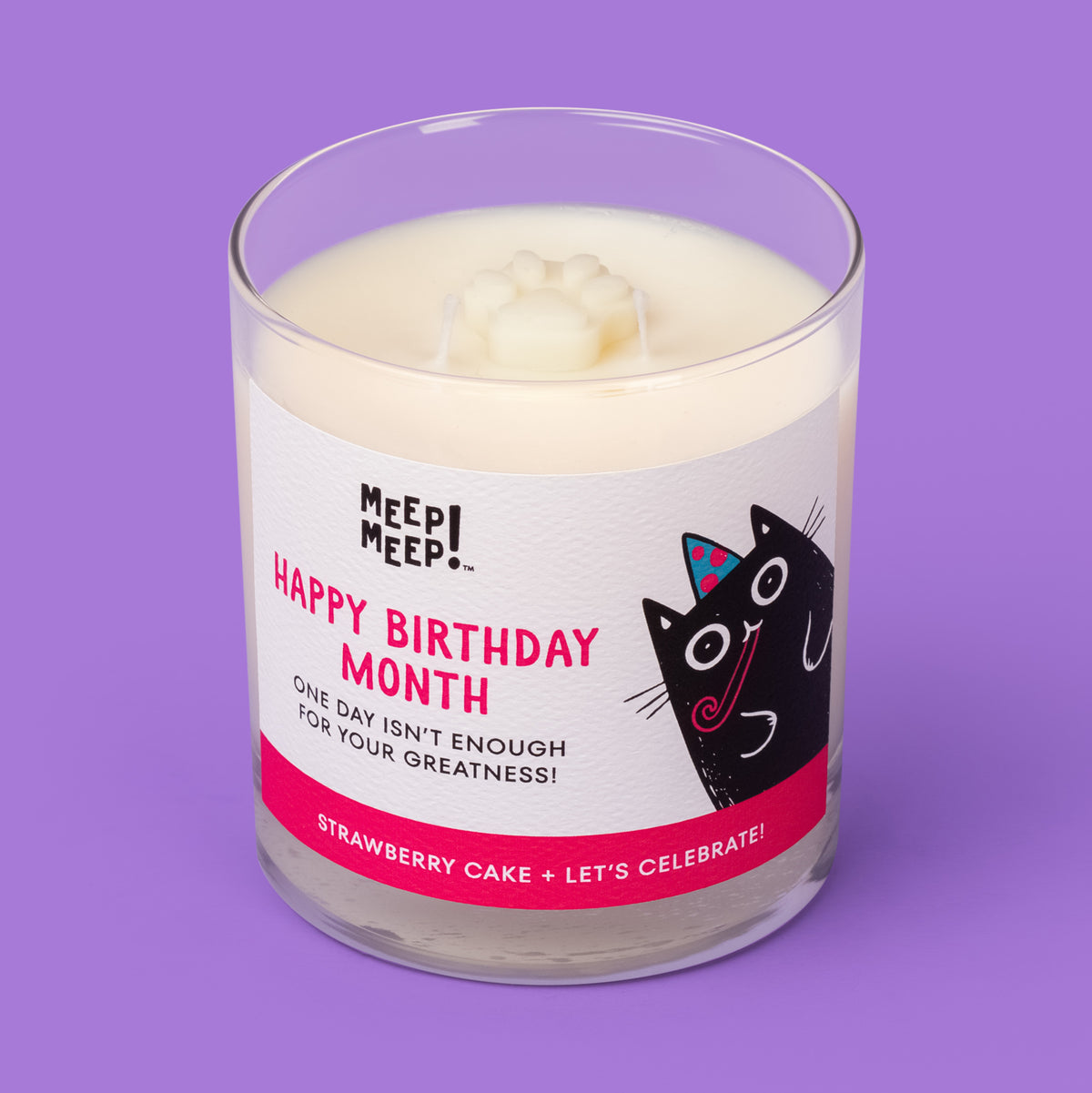 Meep Meep Happy Birthday Month candle in a clear glass jar with a label featuring a black cat wearing a party hat. Jar is angled so we can see the paw embed on top of candle.