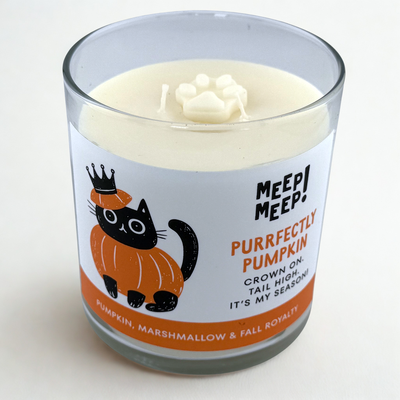 Photograph of Meep Meep candle with wax pawprint embed on top. Label has an Illustration of a cat wearing a pumpkin and crown on head. Text on white background says "Purrfectly Pumpkin, Crown On. Tail High. It's My Season"
Bottom of candle label reads Pumpkin, Marshmallow & Fall Royalty