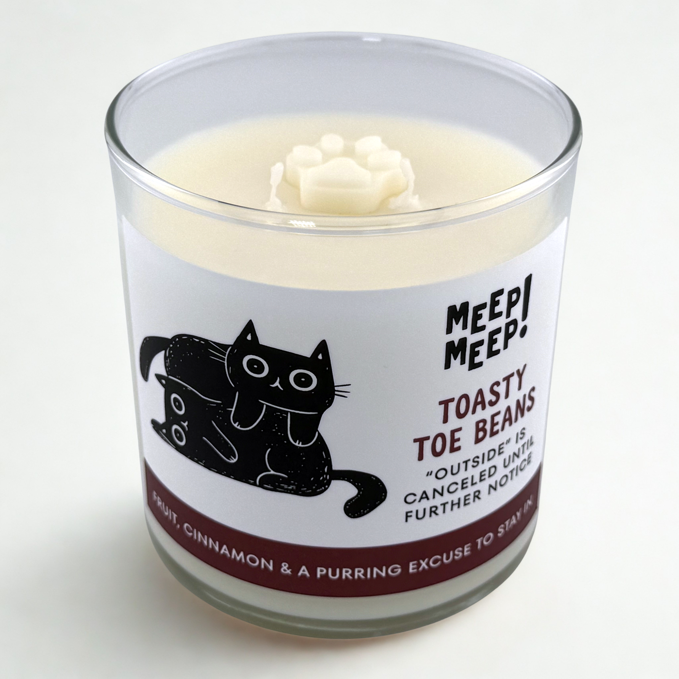 Photograph of Meep Meep  glass candle with wax pawprint embed on top. Label has an Illustration of two cats snuggling. Text on white background says "Toasty Toe Beans. Outside is canceled until further notice"
Bottom of candle label reads fruit, amber & a purring excuse to stay in
