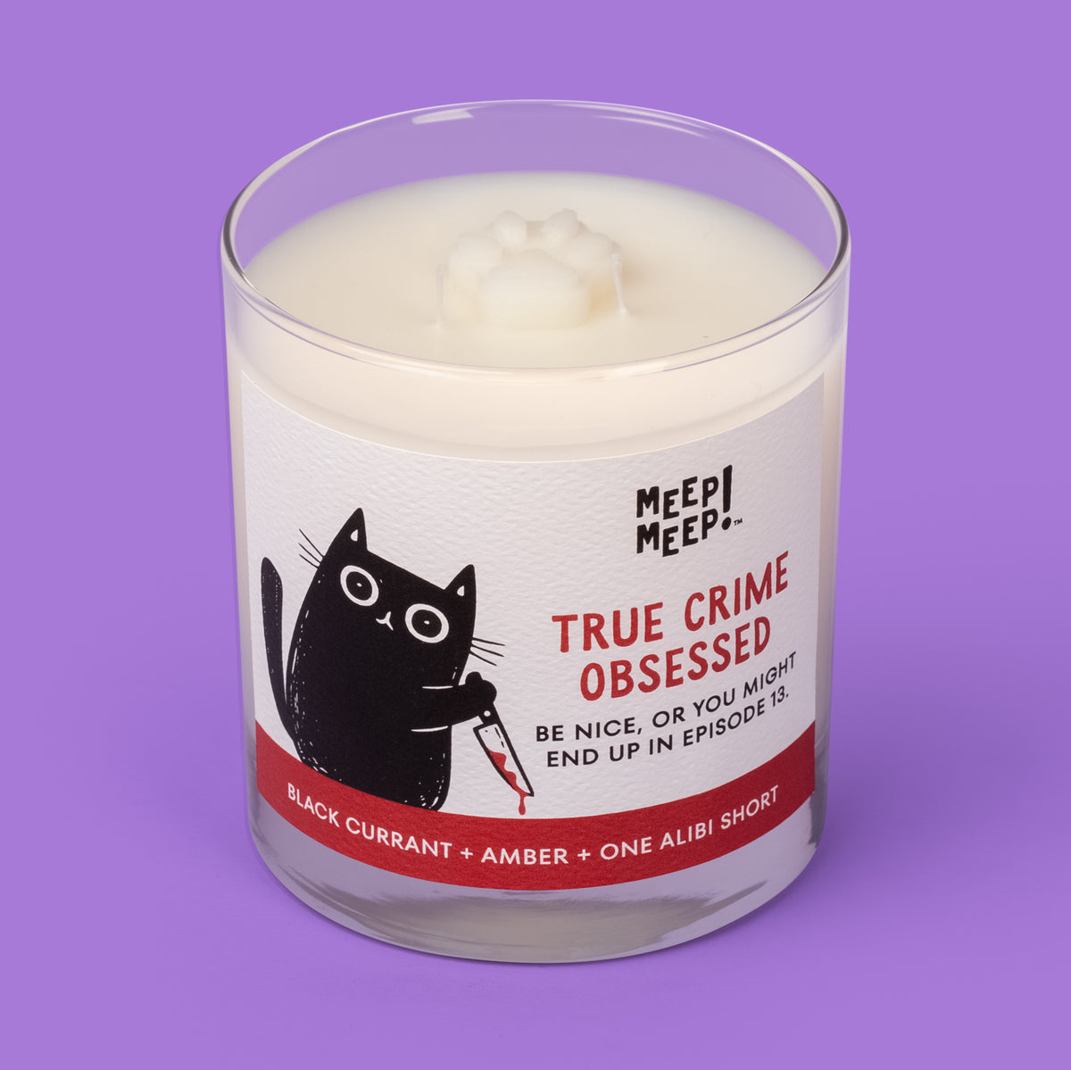 Meep Meep True Crime Obsessed candle in a clear glass jar with a label showing a black cat holding a bloodstained knife. Jar is angled so we can see the paw embed on top of candle.