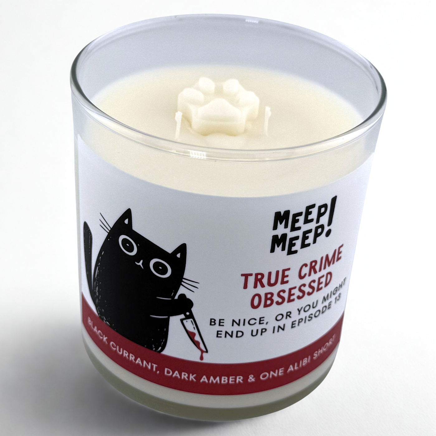 Photograph of Meep Meep candle in glass jar with wax paw print embed on top. Label has an Illustration of a black cat leaning forward holding a bloody knife. Text on white background says "true crimeobSessed. be nice, or you mightend up in episode 13"
Bottom of candle label reads Black Currant, dark amber & one alibi short