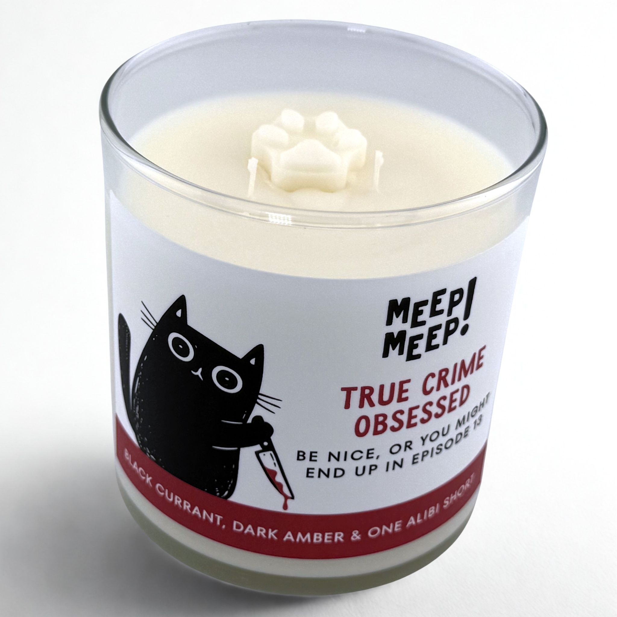 Photograph of Meep Meep candle in glass jar with wax paw print embed on top. Label has an Illustration of a black cat leaning forward holding a bloody knife. Text on white background says "true crimeobSessed. be nice, or you mightend up in episode 13"
Bottom of candle label reads Black Currant, dark amber & one alibi short