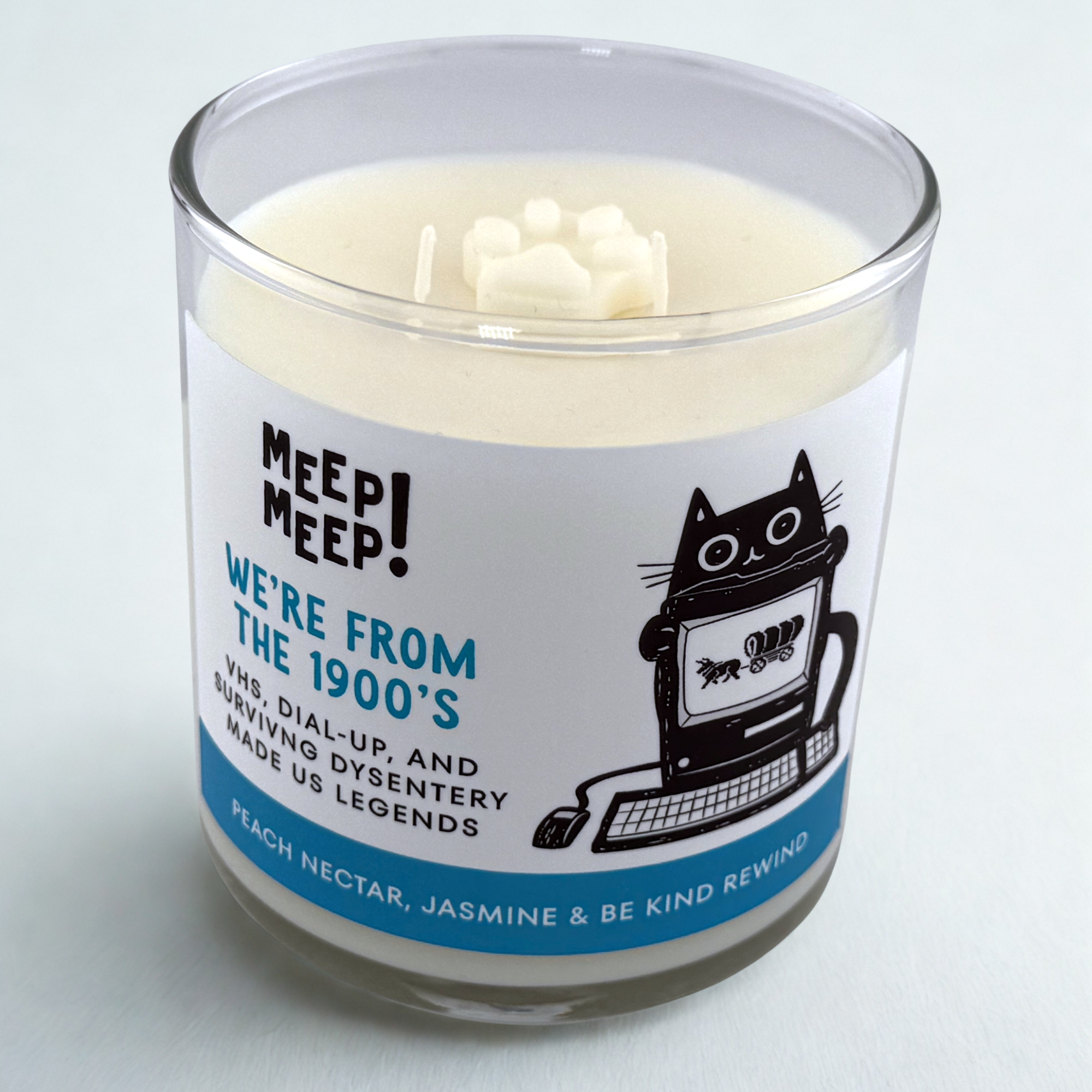 Photograph of Meep Meep candle with wax pawprint embed on top. Label has an Illustration of a cat behind a 1990's computer with an image from the game Oregon Trail on it. Text on white background says "We're from the 1900's. VHS, Dial-up, and surviving dysentery made us legends."
Bottom of candle label reads Peach Nectar, jasmine & be kind rewind