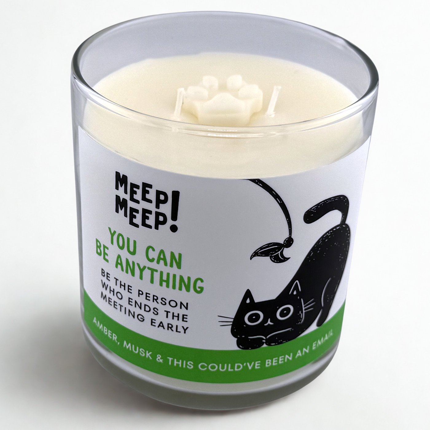 Photograph of Meep Meep candle with wax paw print embed on top. Label has an Illustration of a black cat being playful with a cat toy. Text on white background says "You canbe anything. Be the person who ends the meeting early"
Bottom of candle label reads AMBER, Musk & This Could’ve been an email
