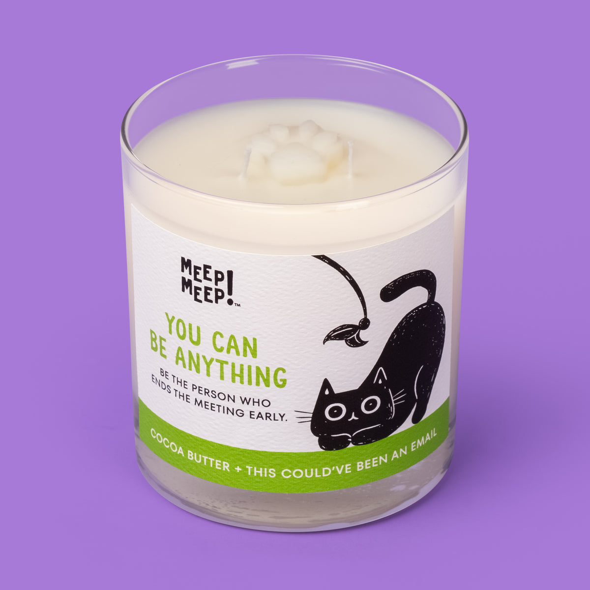 Meep Meep You Can Be Anything candle in a clear glass jar with a green label featuring a black cat crouching beneath a feather toy. Jar is angled so we can see the paw embed on top of candle.