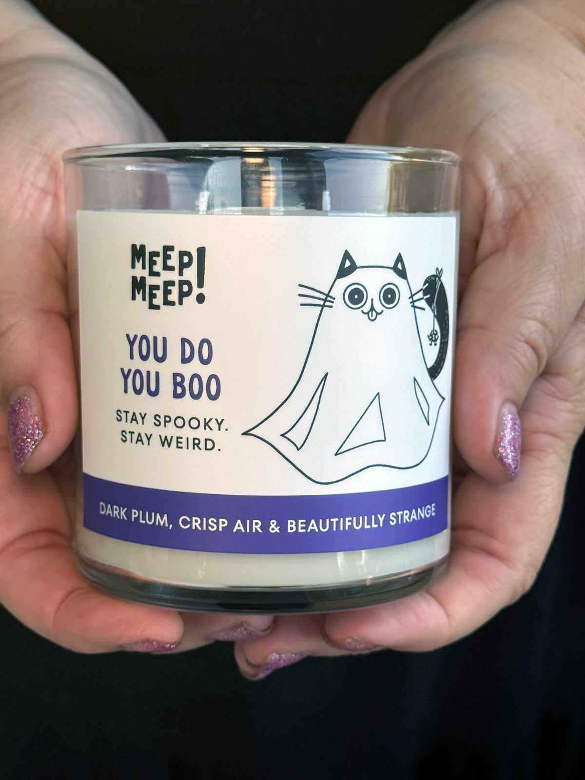 Meep Meep “You Do You Boo” candle in a clear glass jar, featuring a playful ghost cat illustration, purple label accents, and the phrase “Stay Spooky. Stay Weird.” held in two hands.